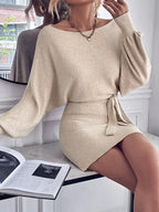 Vestido Curto de Malha com Manga Bufante e Gola Canoa bege, modelo apoiada em bancada mostrando o caimento das mangas amplas