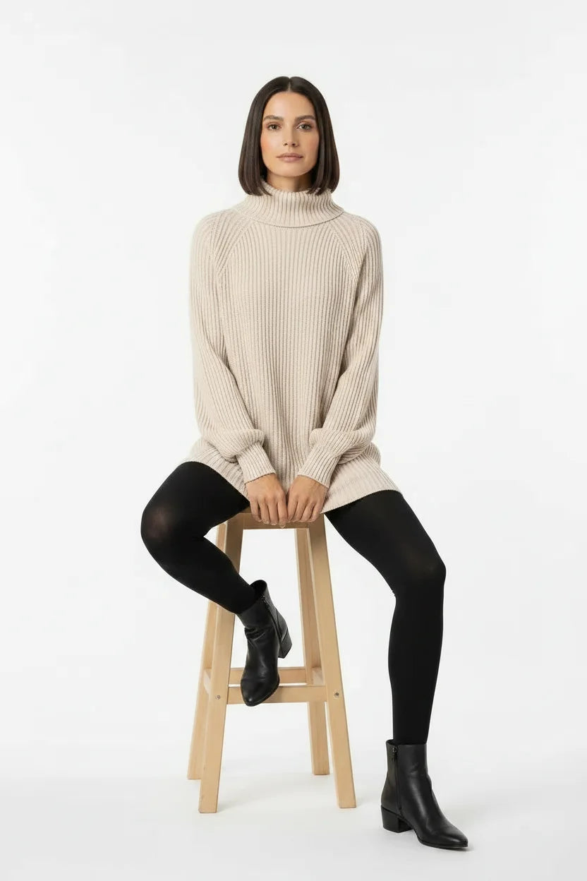 Vestido de Tricot Oversized Gola Alta Bege com a modelo sentada em banqueta, mostrando o caimento amplo da malha
