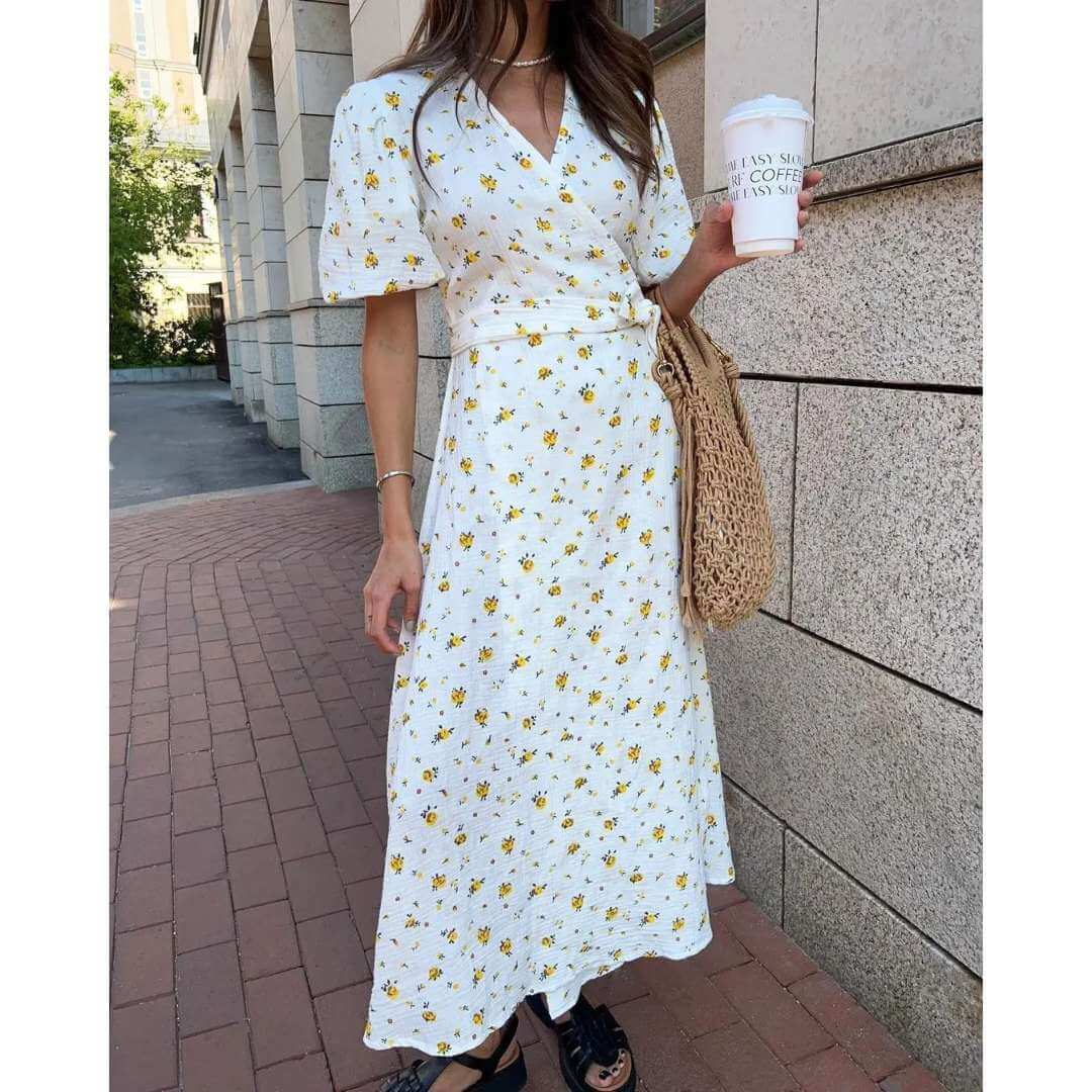 Vestido Feminino Longo Florido Amarelo com a modelo segurando copo de café e bolsa de palha na calçada