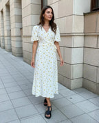 Vestido Feminino Midi Florido Amarelo em cena urbana, visto de frente com faixa na cintura e sandália preta