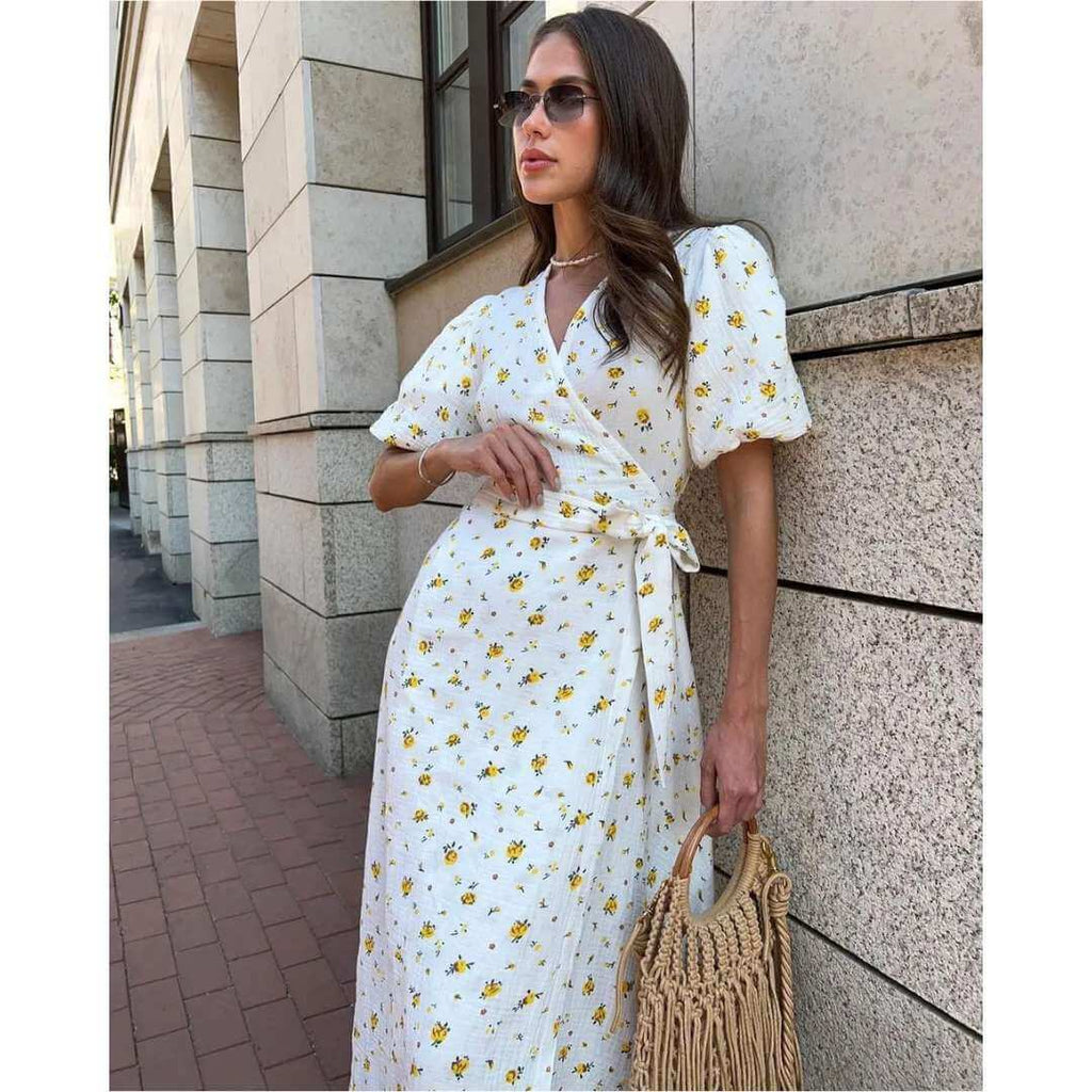 Vestido Feminino Midi Florido Amarelo em pose encostada na parede, com óculos escuros e bolsa de palha